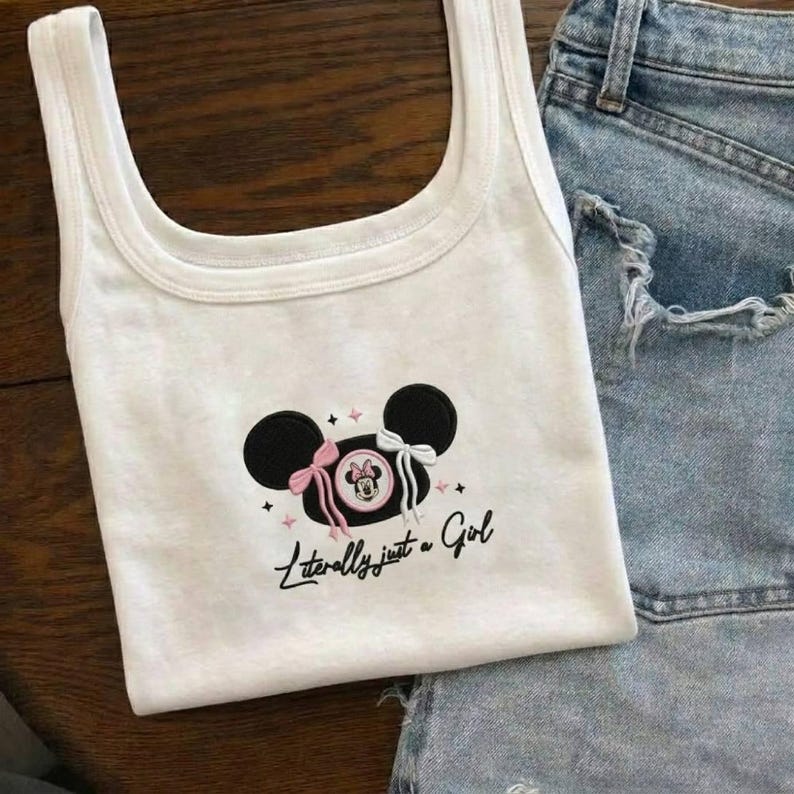 May include: White tank top featuring a Minnie Mouse design with black ears, pink and white bows, and the text "Literally just a Girl." The top is paired with denim shorts, creating a casual, everyday look.
