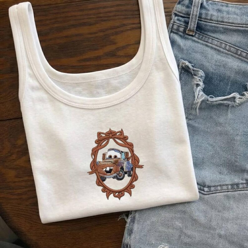 Op de afbeelding: Witte tanktop met een cartoonafbeelding van Takel uit de film Cars, omlijst door een bruine decoratieve rand. De tanktop wordt gecombineerd met een spijkerbroek, wat een casual look cre&euml;ert.