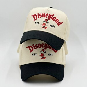 May include: Two beige baseball caps with black brims are stacked. The caps feature the word "Disneyland" in red script, along with a Minnie Mouse graphic and the text "EST. 1955".