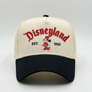 May include: A beige and black baseball cap featuring the word "Disneyland" in red script. The cap also includes the text "EST. 1955" and a Minnie Mouse graphic. The cap has a curved brim and a black button on top.