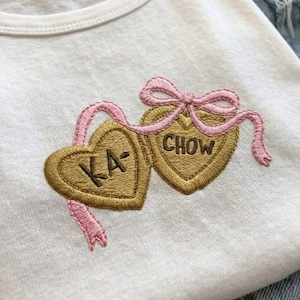 K&ouml;nnte beinhalten: Wei&szlig;es T-Shirt mit einem gestickten Design mit zwei goldfarbenen herzf&ouml;rmigen Anh&auml;ngern. Ein rosa Band und eine Schleife verbinden die Anh&auml;nger. Ein Herz sagt "KA-" und das andere sagt "CHOW".