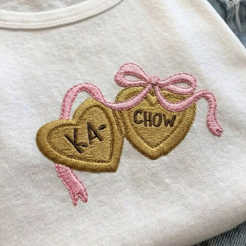 K&ouml;nnte beinhalten: Wei&szlig;es T-Shirt mit einem gestickten Design mit zwei goldfarbenen herzf&ouml;rmigen Anh&auml;ngern. Ein rosa Band und eine Schleife verbinden die Anh&auml;nger. Ein Herz sagt "KA-" und das andere sagt "CHOW".