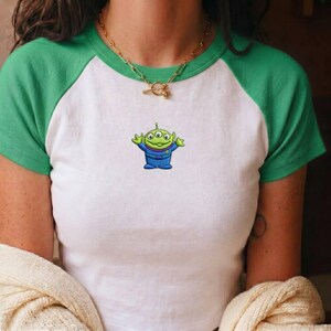 May include: A white and green raglan t-shirt featuring a small embroidered graphic of a three-eyed green alien character wearing a blue suit on the center chest. The shirt has short green sleeves and a matching green crew neckline.