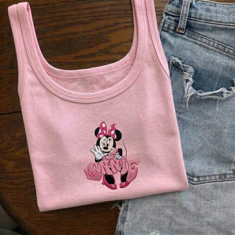 Może przedstawiać: Jasnor&oacute;żowy top bez rękaw&oacute;w z haftowanym wzorem Myszki Minnie. Postać z kresk&oacute;wki ma na sobie r&oacute;żową sukienkę i kokardę. Top bez rękaw&oacute;w jest połączony z dżinsowymi szortami.