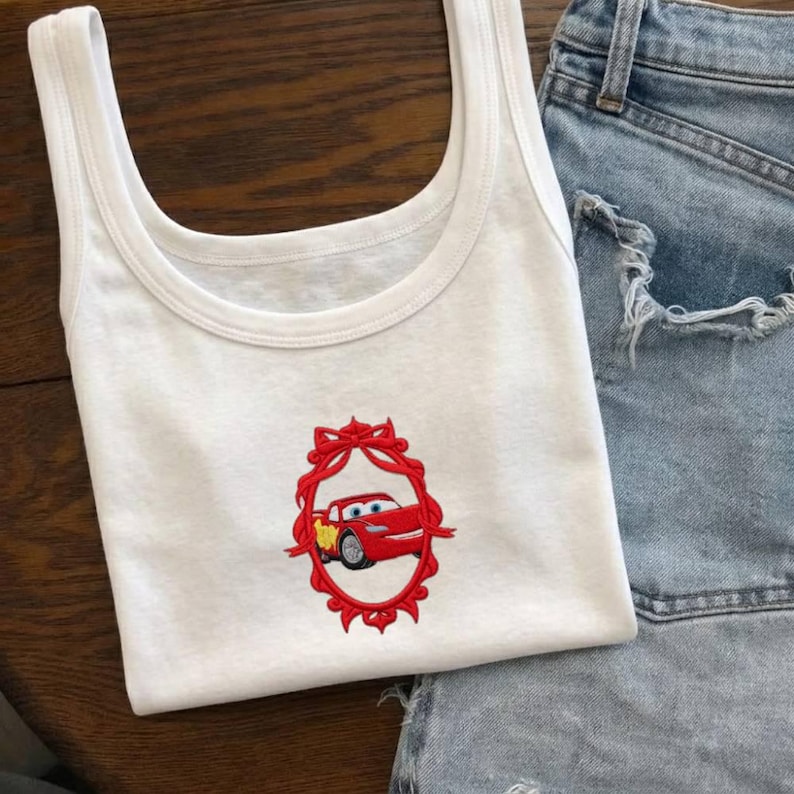 Op de afbeelding: Witte tanktop met een rood geborduurd ontwerp van een cartoon raceauto in een rood decoratief frame. De tanktop wordt gecombineerd met een spijkerbroek. De top heeft een ronde hals en een mouwloos ontwerp.