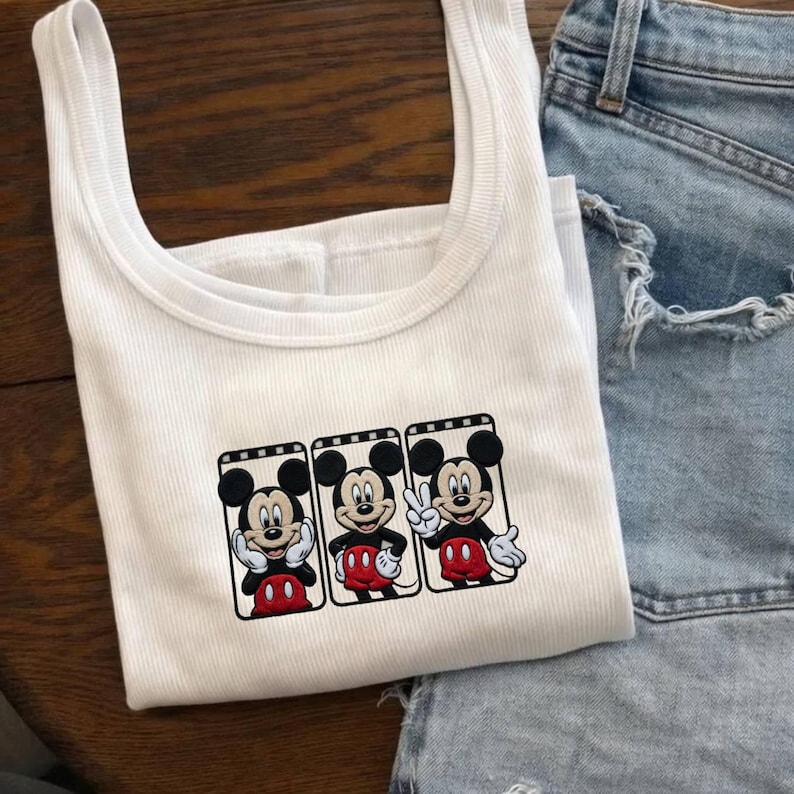 Puede incluir: Camiseta sin mangas blanca acanalada con un gr&aacute;fico de Mickey Mouse en tres poses diferentes. Mickey lleva pantalones cortos rojos y guantes negros. La camiseta sin mangas se combina con pantalones cortos vaqueros azul claro.