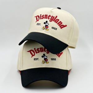 May include: Two beige baseball caps with black brims are stacked. The caps feature the word "Disneyland" in red script, along with a Mickey Mouse graphic and the text "EST. 1955."