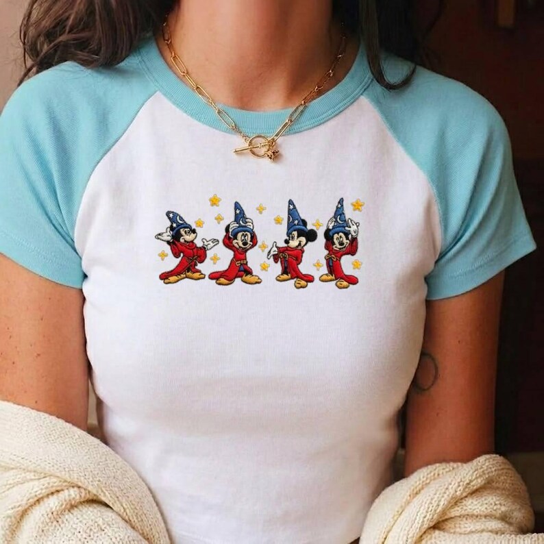May include: A white raglan t-shirt with light blue sleeves featuring a graphic print of four Mickey Mouse characters dressed as wizards in red robes and blue hats, surrounded by small yellow stars, paired with a gold-toned chain link necklace.