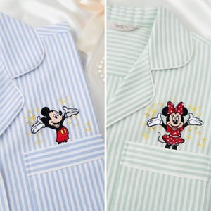 Op de afbeelding: Twee gestreepte pyjamabovenstukken, één lichtblauw en één mintgroen. Het lichtblauwe bovenstuk heeft een geborduurde Mickey Mouse, terwijl het mintgroene bovenstuk een geborduurde Minnie Mouse heeft. Beide hebben een zak.