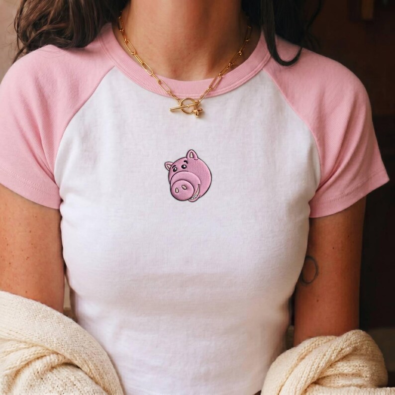 May include: A white and light pink raglan t-shirt featuring a small embroidered pink pig face graphic centered on the chest. The shirt has short sleeves and a round neckline, paired with a gold-toned chain necklace with a toggle clasp.