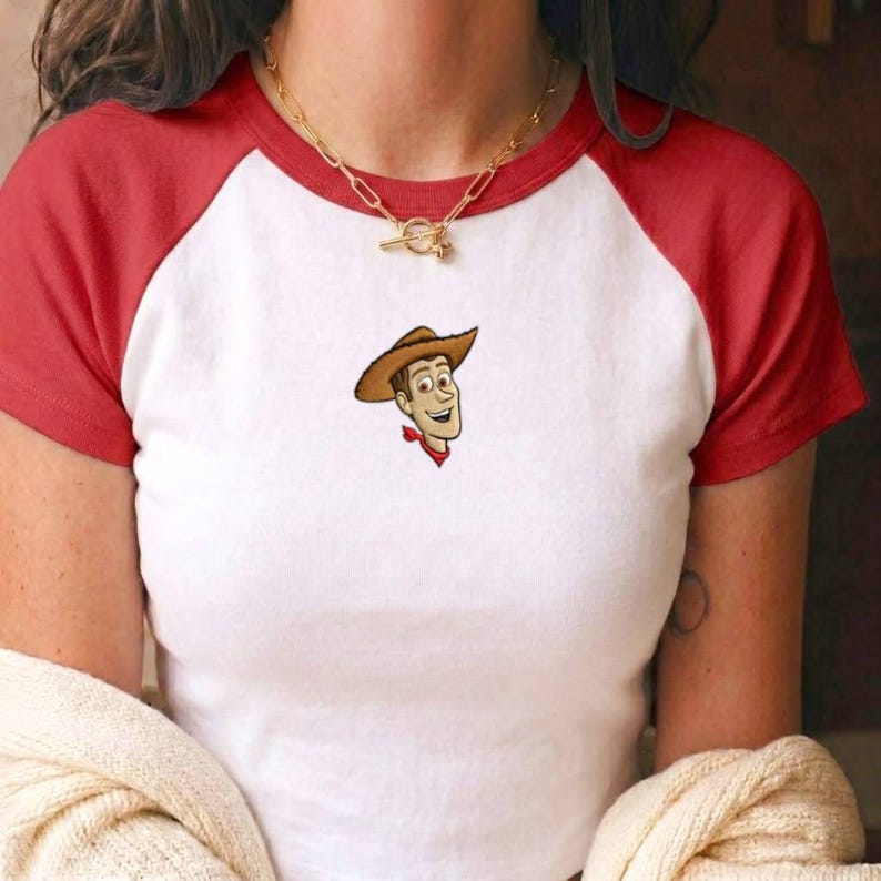 May include: A white raglan t-shirt with red sleeves and a red collar featuring a small embroidered graphic of a cowboy character wearing a brown hat and a red neckerchief centered on the chest, paired with a gold-toned chain link necklace.