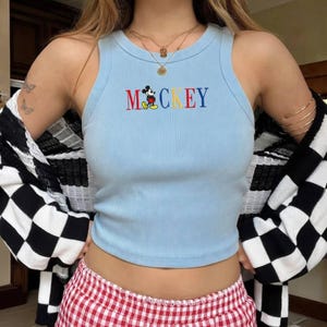Puede incluir: Camiseta sin mangas acanalada azul claro con la palabra "MICKEY" bordada en letras coloridas y una imagen de dibujos animados de Mickey Mouse. La camiseta se combina con pantalones cortos a cuadros rojos y blancos y una rebeca a cuadros blancos y negros.
