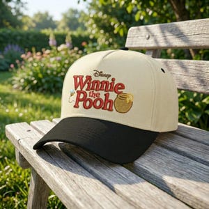 May include: A beige baseball cap with a black brim featuring the text "Disney Winnie the Pooh" in red embroidery, along with a honey pot and bee graphic. The cap is resting on a wooden bench outdoors.
