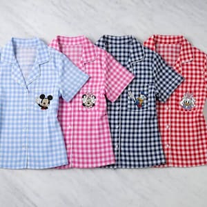 May include: Four short-sleeved pajama shirts in blue, pink, navy, and red gingham patterns. Each shirt features a different Disney character patch: Mickey Mouse, Minnie Mouse, Donald Duck, and Daisy Duck. The shirts have white collars and button-up fronts.