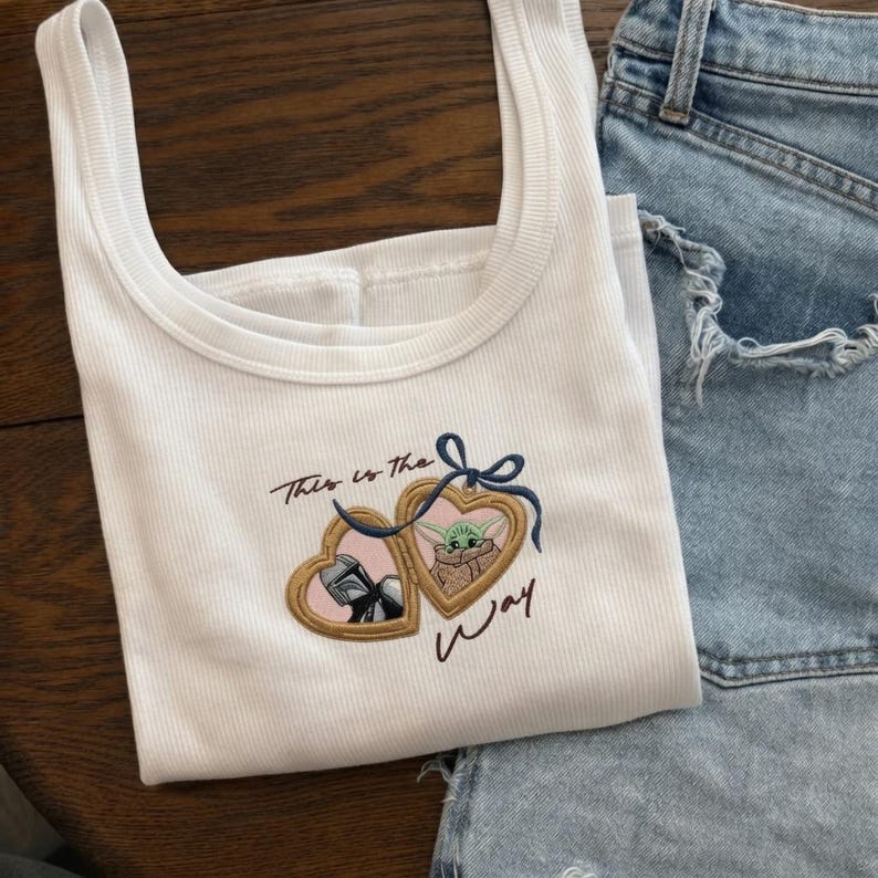 May include: A white ribbed tank top featuring an embroidered design of two gold heart-shaped lockets tied with a blue bow. One locket displays a Mandalorian helmet, the other a green creature, with the text This is the Way embroidered in black.