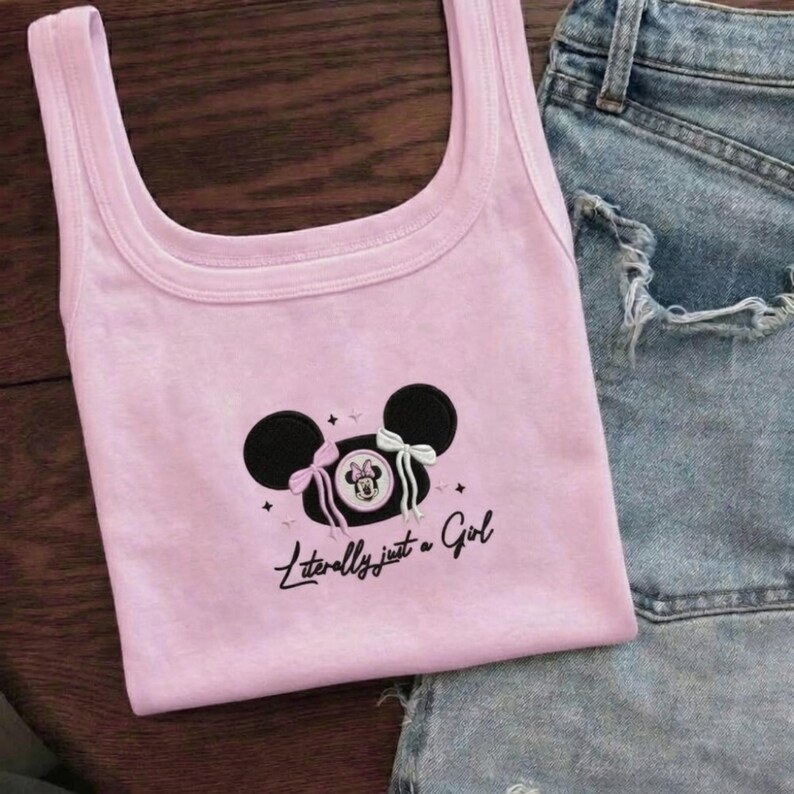 May include: A light pink tank top with a black and white Minnie Mouse design and the text "Literally just a Girl." The tank top is paired with denim shorts. The Minnie Mouse design includes two black circles for ears, a Minnie Mouse image, and bows.