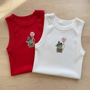 May include: Two sleeveless baby onesies, one red and one white. Each onesie features a cartoon image of a green alien character wearing a wizard hat and holding a pink Mickey Mouse balloon. The onesies are made of a soft, ribbed fabric.