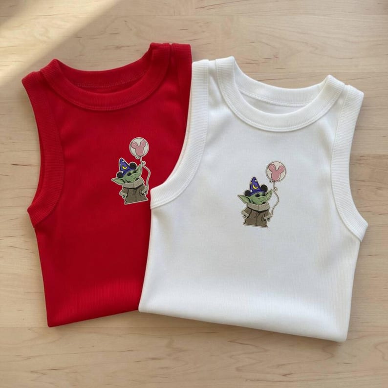 May include: Two sleeveless baby onesies, one red and one white. Each onesie features a cartoon image of a green alien character wearing a wizard hat and holding a pink Mickey Mouse balloon. The onesies are made of a soft, ribbed fabric.