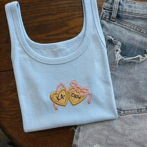 K&ouml;nnte beinhalten: Hellblaues Tanktop mit Rundhalsausschnitt und Stickerei. Das Design zeigt zwei herzf&ouml;rmige Kekse mit den Schriftz&uuml;gen "KA" und "CHOW" in Gold, verbunden durch rosa B&auml;nder und Schleifen. Das Tanktop wird mit Jeansshorts kombiniert.
