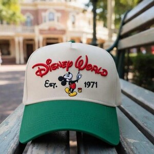 Op de afbeelding: Cr&egrave;mekleurige baseballcap met een groene klep. De cap heeft de woorden "Disney World" in het rood, de afbeelding van Mickey Mouse en de tekst "Est. 1971". De cap ligt op een houten bank.