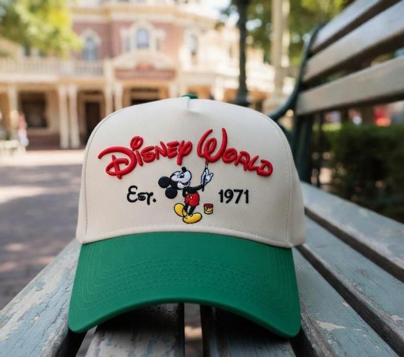 Op de afbeelding: Cr&egrave;mekleurige baseballcap met een groene klep. De cap heeft de woorden "Disney World" in het rood, de afbeelding van Mickey Mouse en de tekst "Est. 1971". De cap ligt op een houten bank.