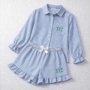 Puede incluir: Conjunto de pijama a rayas azules y blancas. La parte superior de manga larga tiene una botonadura y un bolsillo en el pecho con las letras "XYZ" bordadas. Los pantalones cortos tienen un dobladillo con volantes y un lazo blanco. El conjunto es de tela suave y ligera.