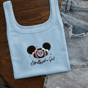 May include: A light blue tank top with a black Mickey Mouse design, pink and white bows, and the text "Literally just a Girl". The tank top is paired with denim shorts.