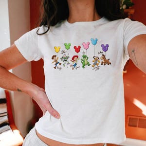 May include: White t-shirt featuring a colorful graphic of Toy Story characters holding Mickey Mouse-shaped balloons. The characters include Woody, Buzz Lightyear, Jessie, Rex, Slinky Dog, and Bullseye. Each character has a signature below.
