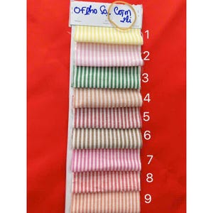 May include: A fabric swatch card featuring nine striped fabric samples in various colors, including yellow, pink, green, and brown. The samples are arranged vertically, with a label at the top that reads "Of pho So.. Com li".