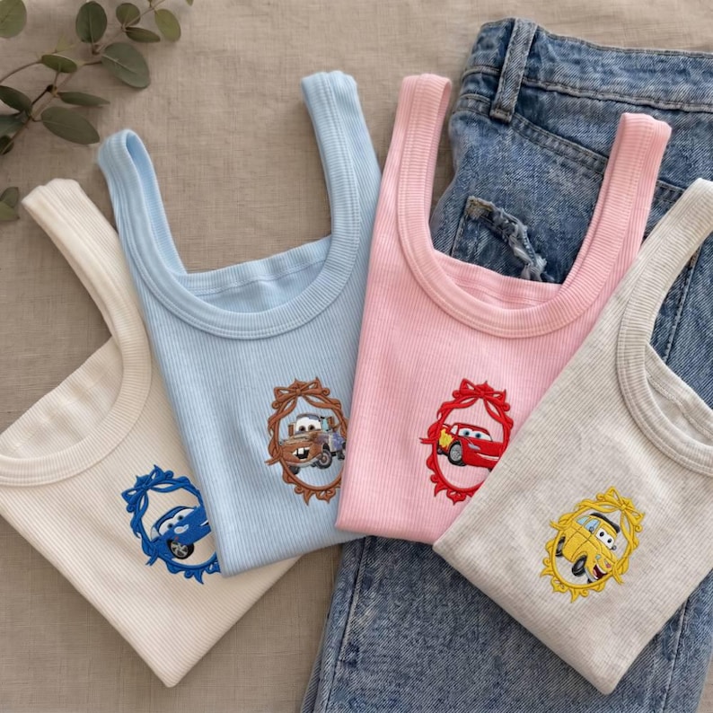 Op de afbeelding: Vier geribbelde tanktops in cr&egrave;me, lichtblauw, roze en grijs, elk geborduurd met een cartoon auto-ontwerp in een decoratief frame. De tanktops worden getoond met een spijkerbroek.