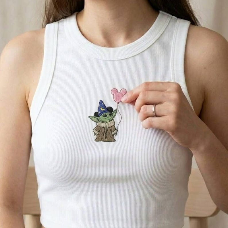 May include: White ribbed tank top featuring an embroidered design of a green alien character wearing a wizard hat and holding a pink Mickey Mouse balloon. The character is wearing a brown coat.