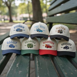 Op de afbeelding: Een verzameling baseballpetten met verschillende auto-ontwerpen en namen als "Hudson", "Sally", "The King", "Mater", "Fillmore", "McQueen" en "Sheriff". Elke pet heeft een ander kleurenschema, met tekst en auto-illustraties.