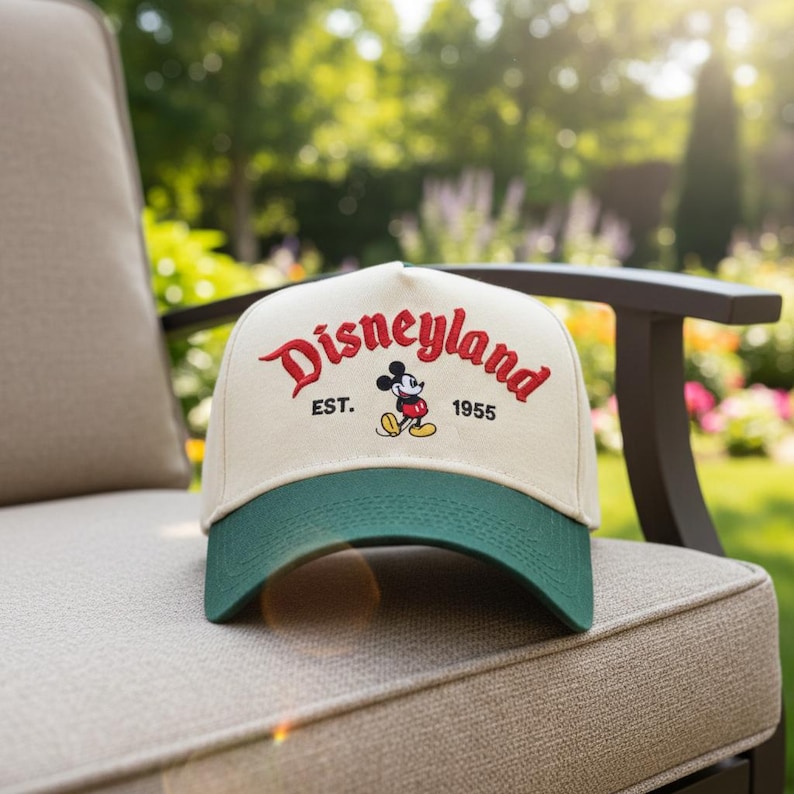 May include: A beige and green baseball cap with the word "Disneyland" in red script. The cap features an image of Mickey Mouse and the text "EST. 1955". The hat is resting on a beige outdoor chair.