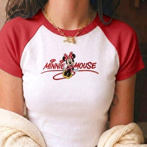 Op de afbeelding: Een wit raglan-t-shirt met rode mouwen en een rode kraag, voorzien van een geborduurde afbeelding van Minnie Mouse en de rode tekst "MINNIE MOUSE" op de borst. Om de hals wordt een goudkleurige ketting met een knevelsluiting gedragen.