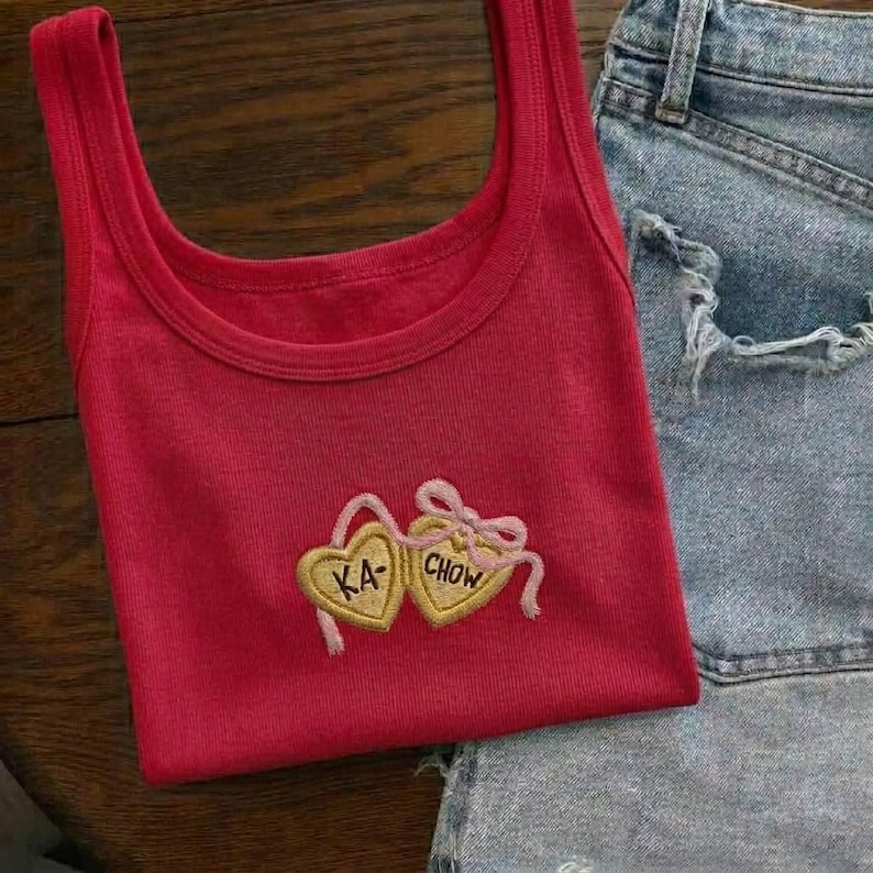 K&ouml;nnte beinhalten: Ein rotes Tanktop mit Rundhalsausschnitt und einem herzf&ouml;rmigen Stickdesign. Das Design zeigt zwei goldene Herzen mit den Texten "KA" und "CHOW" und eine rosa Schleife. Das Tanktop wird mit Jeansshorts kombiniert.