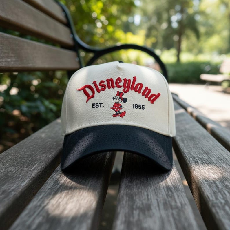 May include: A beige and navy blue baseball cap with the word "Disneyland" in red script. The cap features a Minnie Mouse graphic and the text "EST. 1955". The cap is sitting on a wooden bench.