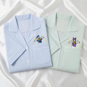 May include: Two pajama tops, one light blue and one light green, both with vertical stripes. Each has a pocket with an embroidered cartoon character: Donald Duck and Daisy Duck. The pajamas are folded on a white satin surface.