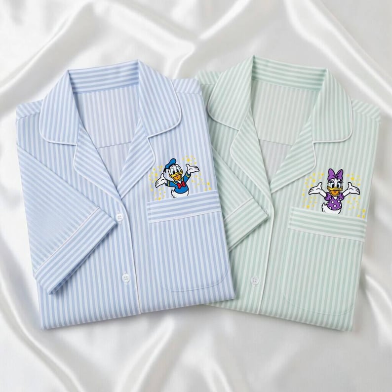 May include: Two pajama tops, one light blue and one light green, both with vertical stripes. Each has a pocket with an embroidered cartoon character: Donald Duck and Daisy Duck. The pajamas are folded on a white satin surface.