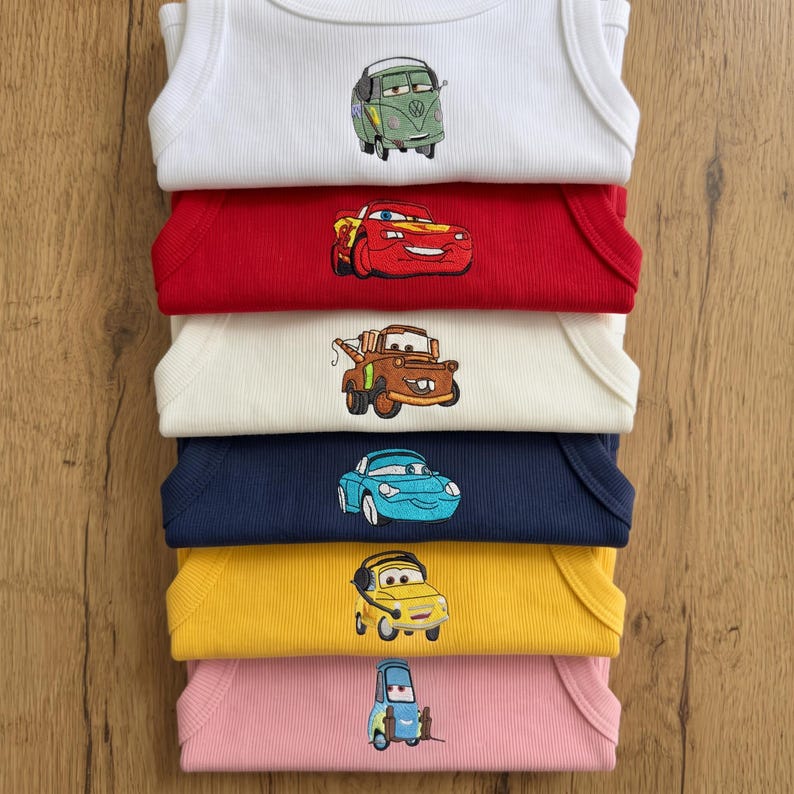 May include: A stack of six baby onesies in white, red, cream, navy, yellow, and pink. Each onesie features an embroidered cartoon car design. The cars are in various colors and styles, including a green van, a red race car, and a brown tow truck.