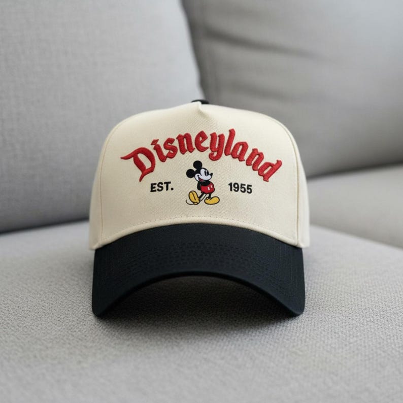 May include: A cream-colored baseball cap with a black brim. The cap features the word "Disneyland" in red script, along with "EST. 1955" and a Mickey Mouse graphic.