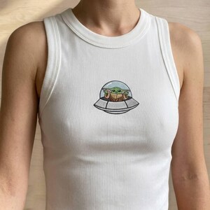 K&ouml;nnte beinhalten: Wei&szlig;es geripptes Tanktop mit Rundhalsausschnitt. Das Tanktop zeigt ein gesticktes Bild eines gr&uuml;nen Alien-Charakters in einem Raumschiff. Das Raumschiff ist grau mit einer transparenten Kuppel. Der Au&szlig;erirdische hat gro&szlig;e Augen und spitze Ohren.