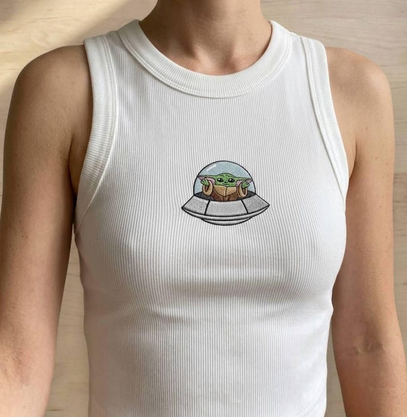 K&ouml;nnte beinhalten: Wei&szlig;es geripptes Tanktop mit Rundhalsausschnitt. Das Tanktop zeigt ein gesticktes Bild eines gr&uuml;nen Alien-Charakters in einem Raumschiff. Das Raumschiff ist grau mit einer transparenten Kuppel. Der Au&szlig;erirdische hat gro&szlig;e Augen und spitze Ohren.