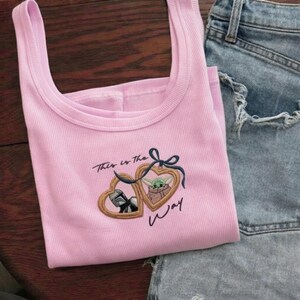May include: A light pink ribbed tank top featuring an embroidered design of two heart-shaped lockets tied with a dark blue bow. One locket displays a Mandalorian helmet, the other a green creature, with the text This is the way embroidered below.