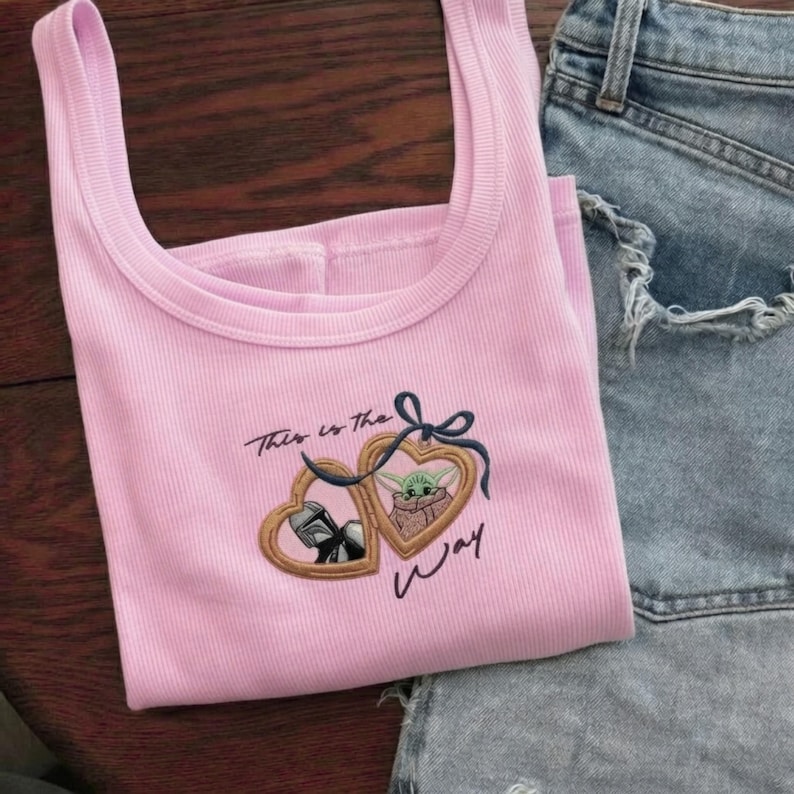 May include: A light pink ribbed tank top featuring an embroidered design of two heart-shaped lockets tied with a dark blue bow. One locket displays a Mandalorian helmet, the other a green creature, with the text This is the way embroidered below.