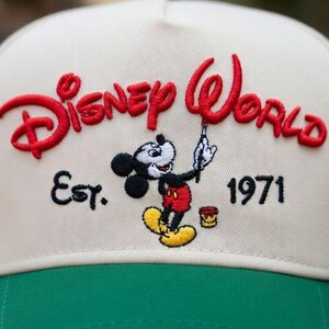 Op de afbeelding: Cr&egrave;me en groene baseballcap met het geborduurde opschrift "Disney World" in rood, Mickey Mouse en het jaar "1971". De cap heeft een klassiek ontwerp.