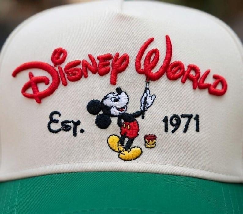 Op de afbeelding: Cr&egrave;me en groene baseballcap met het geborduurde opschrift "Disney World" in rood, Mickey Mouse en het jaar "1971". De cap heeft een klassiek ontwerp.