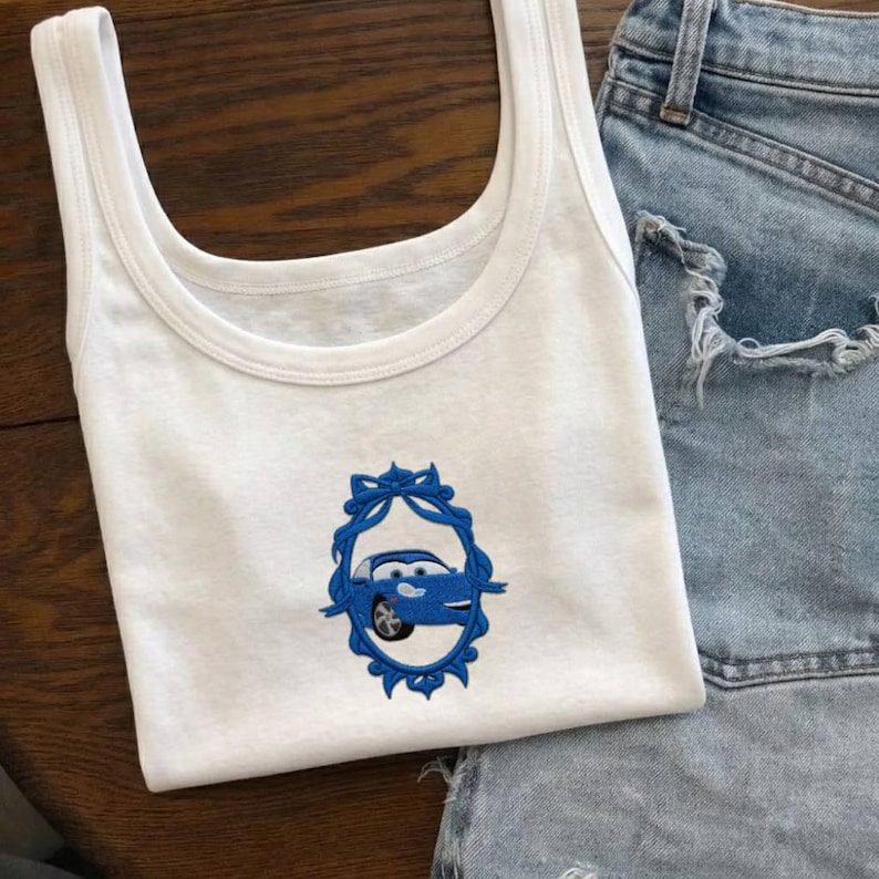 Op de afbeelding: Witte tanktop met een blauw geborduurd autodesign in een decoratief kader. De tanktop wordt getoond met een spijkerbroek. Het autodesign is een cartoon illustratie.