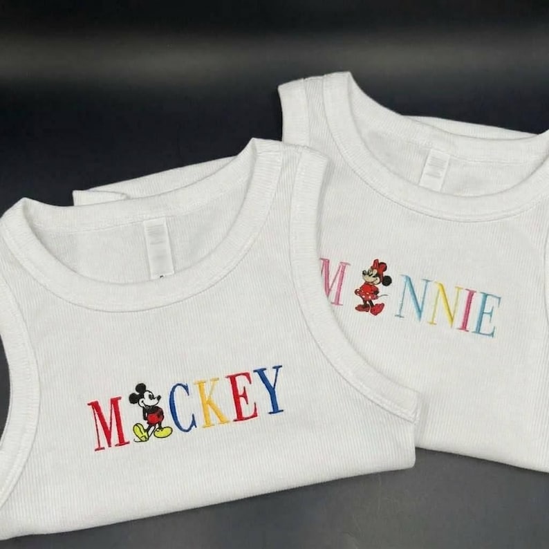 Puede incluir: Dos camisetas sin mangas blancas de canal&eacute; con texto bordado y personajes de dibujos animados. Una camiseta dice "Mickey" en rojo, azul, amarillo y negro, con un rat&oacute;n de dibujos animados. La otra dice "Minnie" en rosa, azul y amarillo, con un rat&oacute;n de dibujos animados.