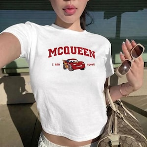 May include: White crop top featuring the word "MCQUEEN" in red, a cartoon image of a red race car, and the words "i am speed". The top is a short-sleeved style, suitable for casual wear.
