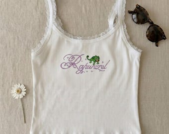 Op de afbeelding: Witte geribbelde tanktop met kanten rand op de bandjes. De voorkant is voorzien van het geborduurde woord "Rapunzel" in paars schrift, samen met een groene kameleon illustratie. Schildpad zonnebrillen zijn rechts geplaatst.
